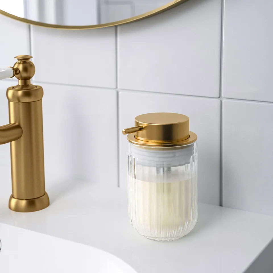 A clear glass soap dispenser with a brass pump sits on a white sink, reflecting a gold-framed round mirror on a white tile wall.