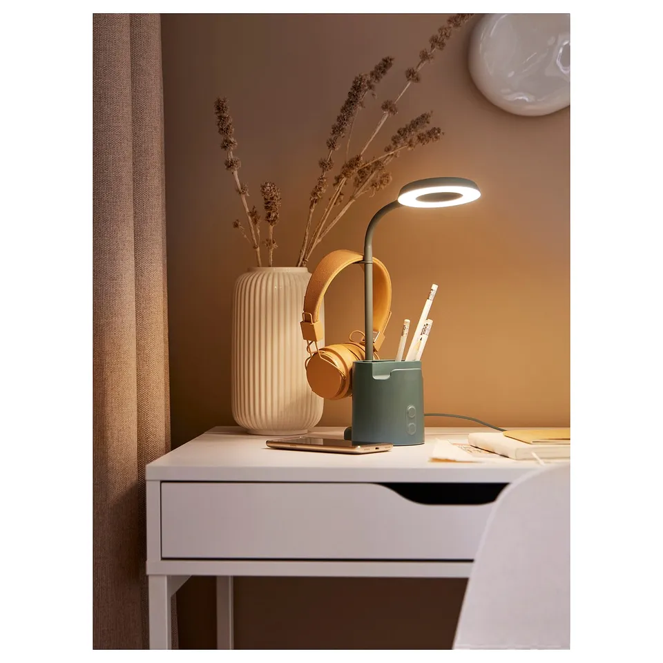 White desk with teal BRUNBÅGE lamp, pen holder, and dried flowers. Headphones hang from hook.