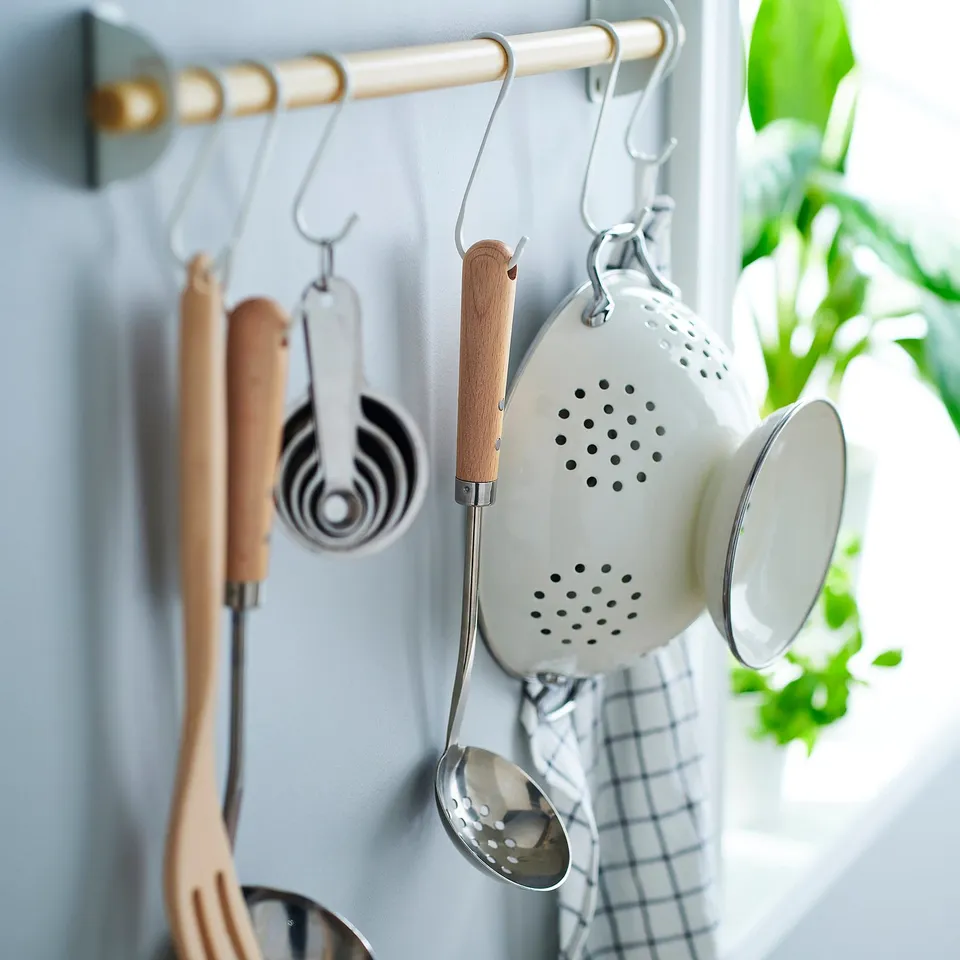 Wall-mounted kitchen utensils: skimmer, ladle, spatula, wooden handles. Blue background with greenery.