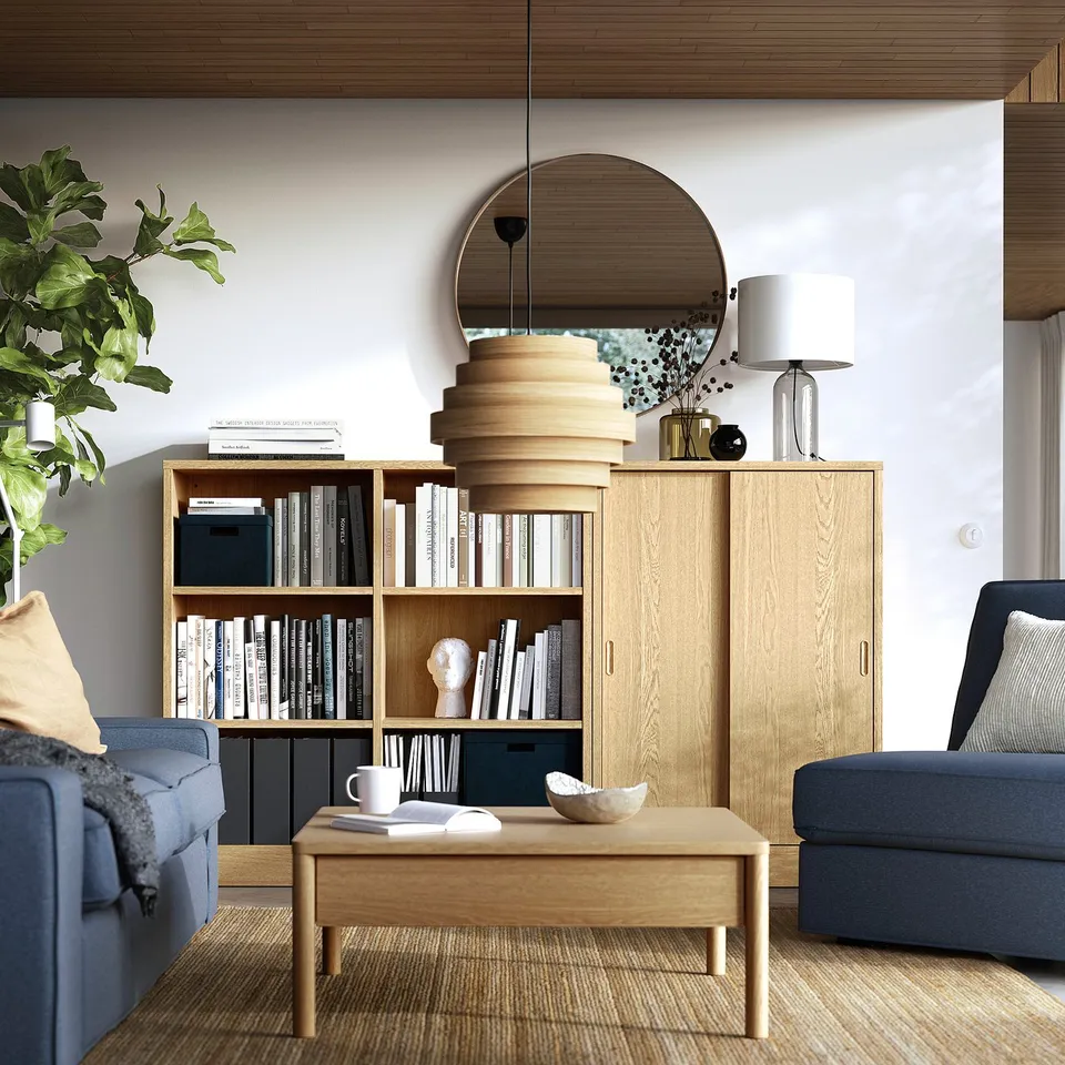 Modern living room with light wood TONSTAD bookcase, mirror, blue couch, plants, and coffee table.
