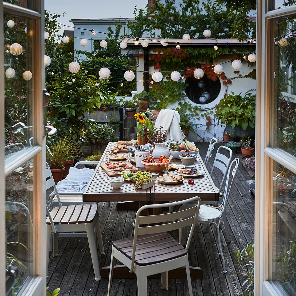 Outdoor dining setup on wooden deck, featuring metal table with food, surrounded by NORRMANSÖ chairs. Decorated with string lights and potted plants.