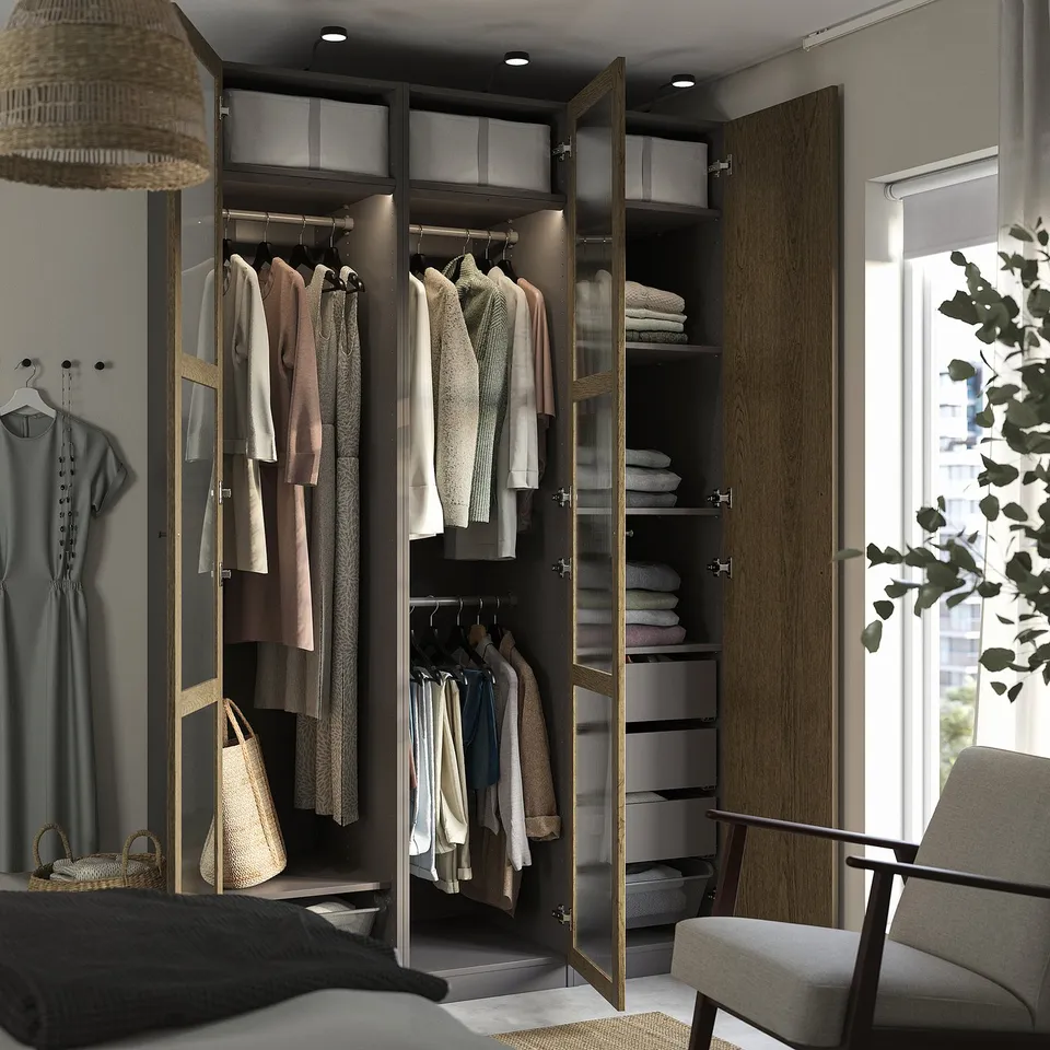 Modern bedroom with open wardrobe holding clothes, folded towels, and baskets. Lighting inside, grey upholstered chair nearby.